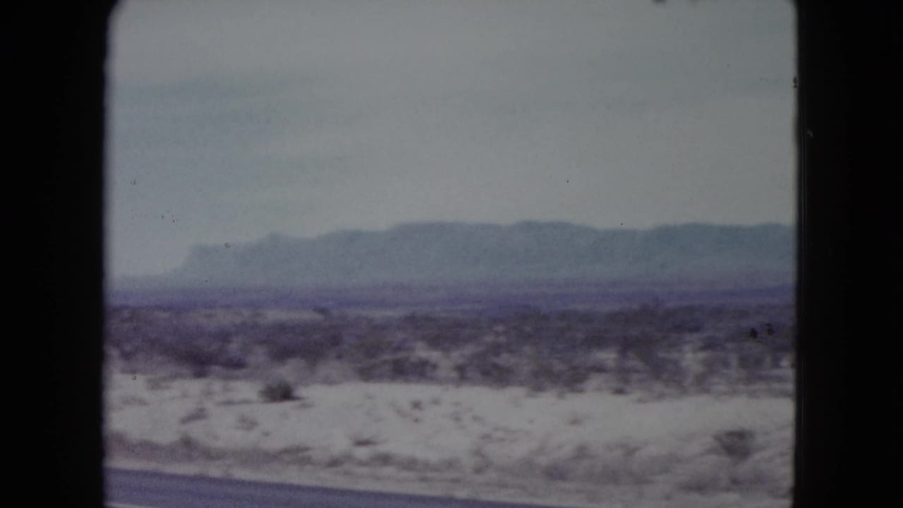Desert scrubland with distant mountains under a pale sky, framed by dark film borders.
