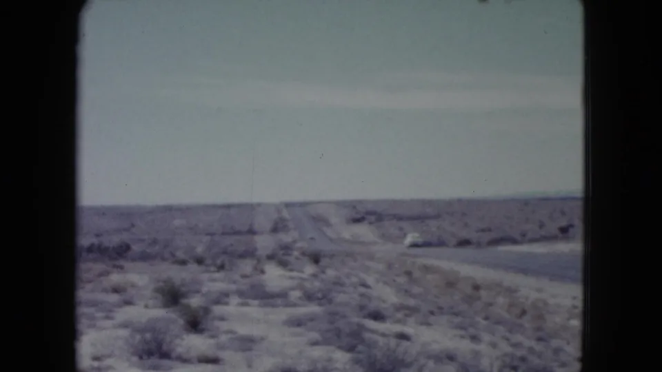 1960 Nevada 8mm frame showing a long rural road through desert scrub with a small white car far ahead.