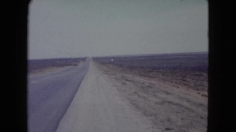 1960 Nevada 8mm frame with a long paved road cutting through open desert scrub and a broad, pale sky.
