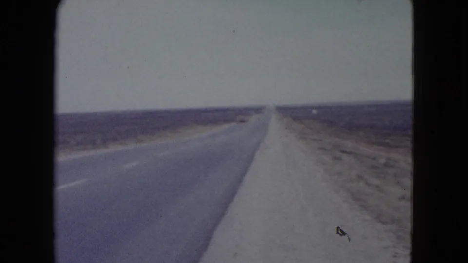 1960 Nevada 8mm frame showing the road edge, pale shoulder, flat scrubland, and a straight route fading into the horizon.