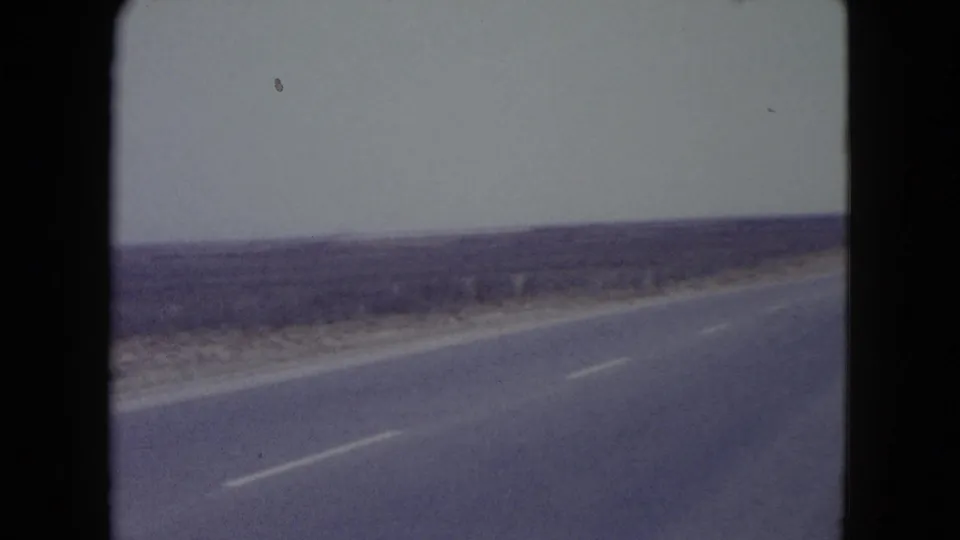1960 Nevada 8mm frame with the paved road crossing the foreground beside open desert, low brush, and a pale horizon.