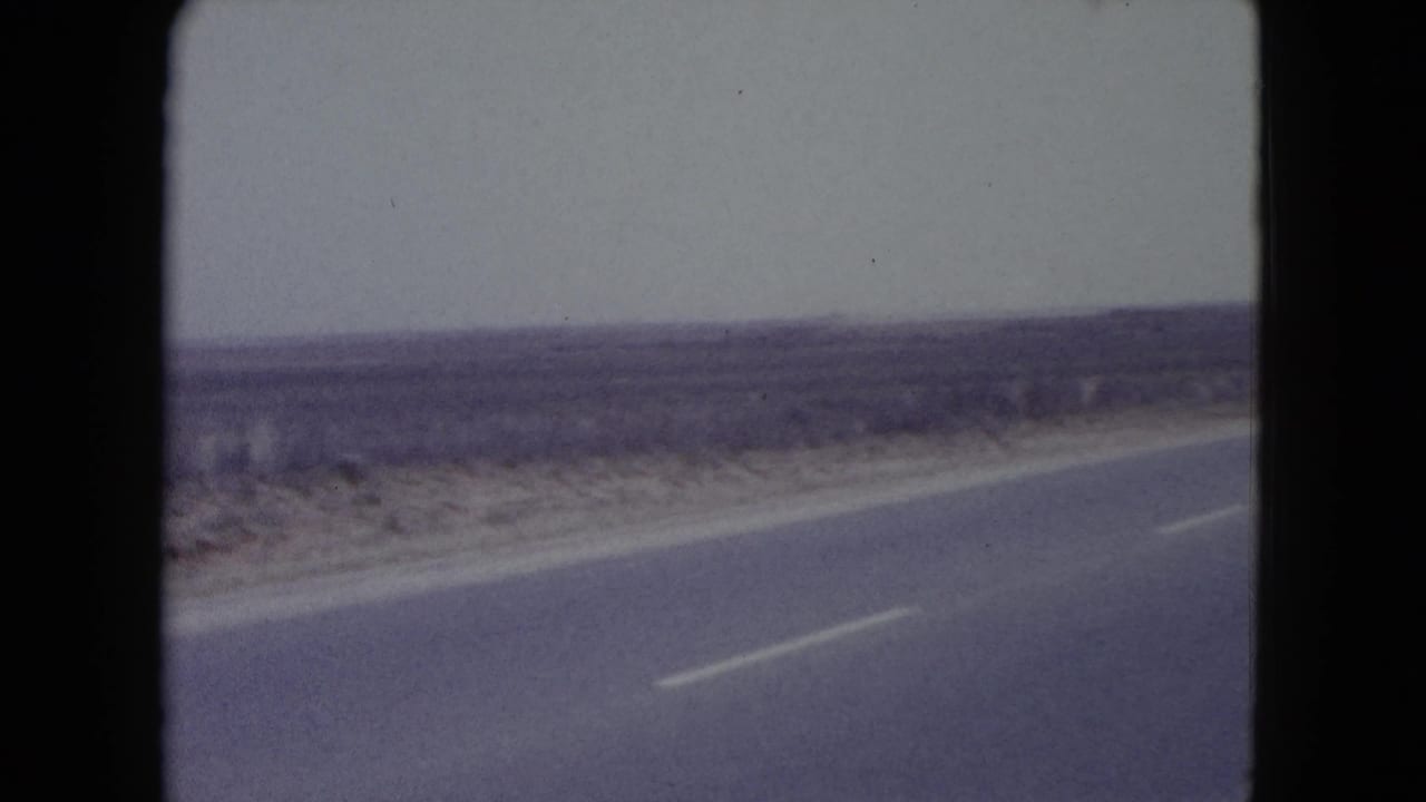 Paved desert road seen from a moving vehicle with scrubland and pale sky.