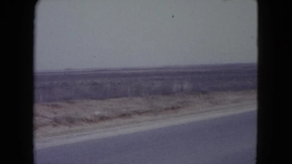 1960 Nevada desert road view from a vehicle, with asphalt edge, dry shoulder, and sparse scrub stretching outward.