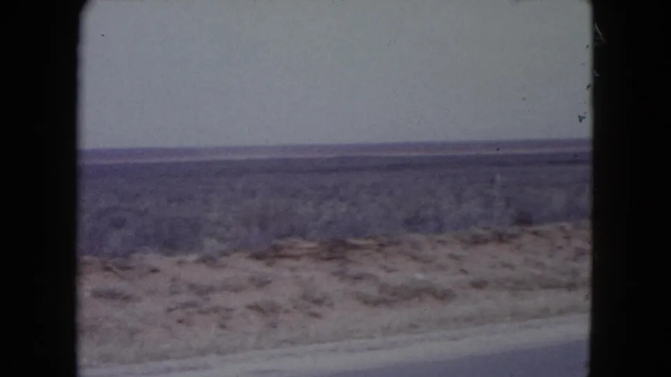 1960 Nevada home-movie still showing open desert scrubland beyond a paved road edge and pale sky.