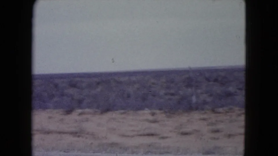 Faded 8mm frame of dry Nevada terrain, likely filmed from a moving traveler perspective, with scrub and a low horizon.
