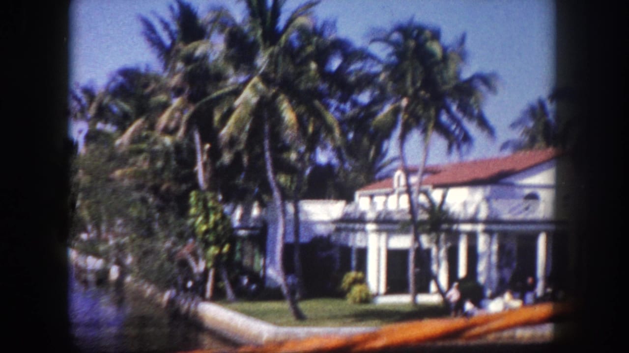 Boat-view frame of a palm-lined waterfront home with a white facade, red tile roof, lawn, and wooden rail in front.
