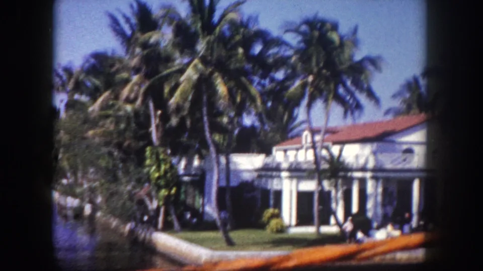 1960 Tampa home-movie frame from a boat passing a white waterfront residence with palms, lawn, red tile roof, and a wooden rail in the foreground.