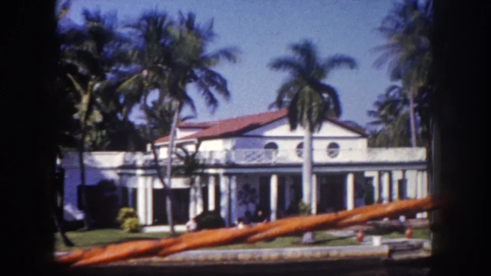 1960 Tampa boat-ride view of a white Mediterranean-style waterfront house, palm trees, lawn, columns, and a rail across the foreground.