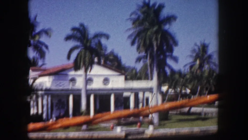 1960 Tampa frame showing a close pass by a white waterfront residence with circular windows, columns, palms, lawn, and boat rail.