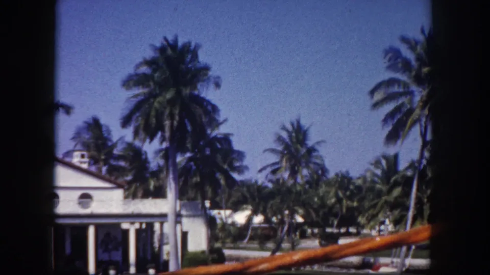 1960 Tampa home-movie frame with a partial white waterfront house, tall palms, lawn, blue sky, and boat rail from a moving vantage point.