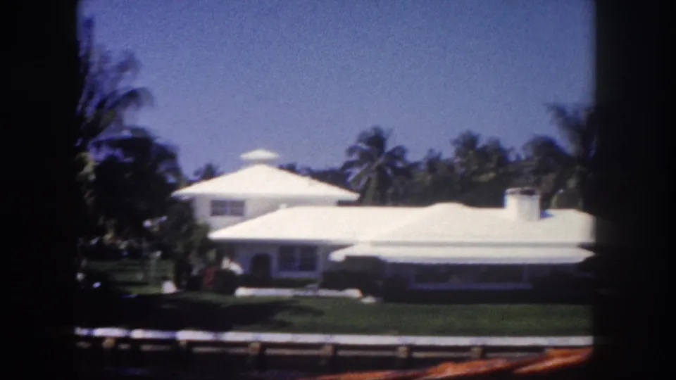 1960 Tampa home-movie frame of a large white residence with a bright roof, lawn, palms, and soft daylight exposure.