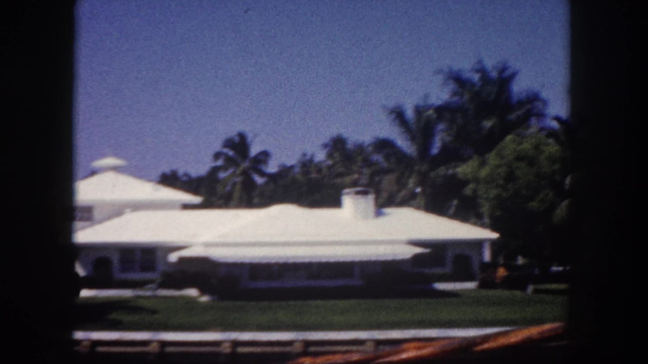 White waterfront house with a pale roof, lawn, palm trees, and dark film borders.