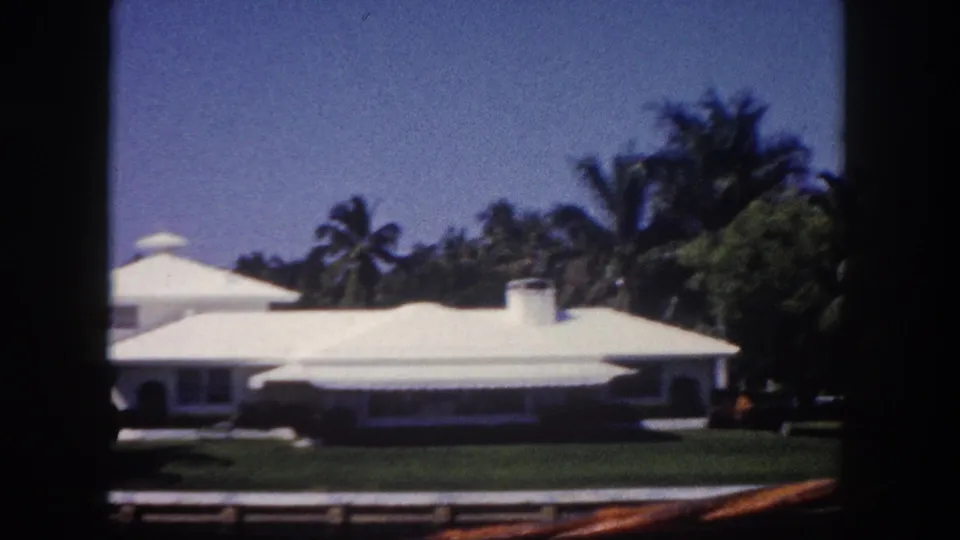 1960 Tampa home-movie frame showing a white waterfront residence, green lawn, palm trees, and soft 8mm edge shadows.