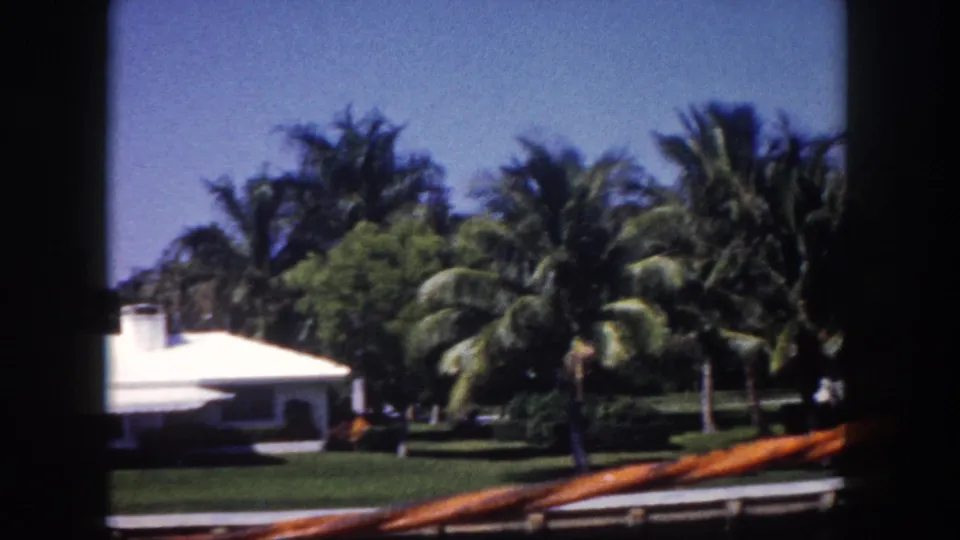 1960 Tampa waterfront view of a white home and palm-lined lawn, partly obscured by dark 8mm frame edges.