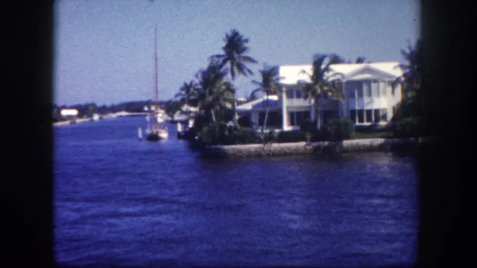1960 Tampa waterfront frame with a white house, palms, seawall, blue water, and a small sailboat near the shore.