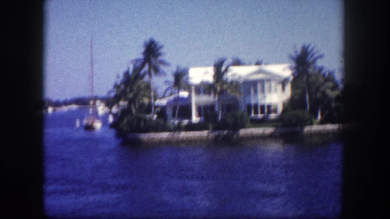 White waterfront house with palms beside blue water and a small sailboat nearby.