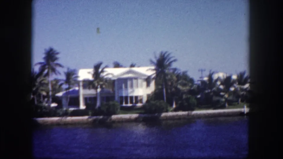 1960 Tampa waterfront view with a large white residence, palms, seawall, and calm blue canal water in soft 8mm color.