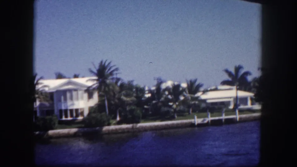 1960 Tampa frame showing a white canal-side home, neighboring residence, palm trees, seawall, and water in a home-movie pass-by.