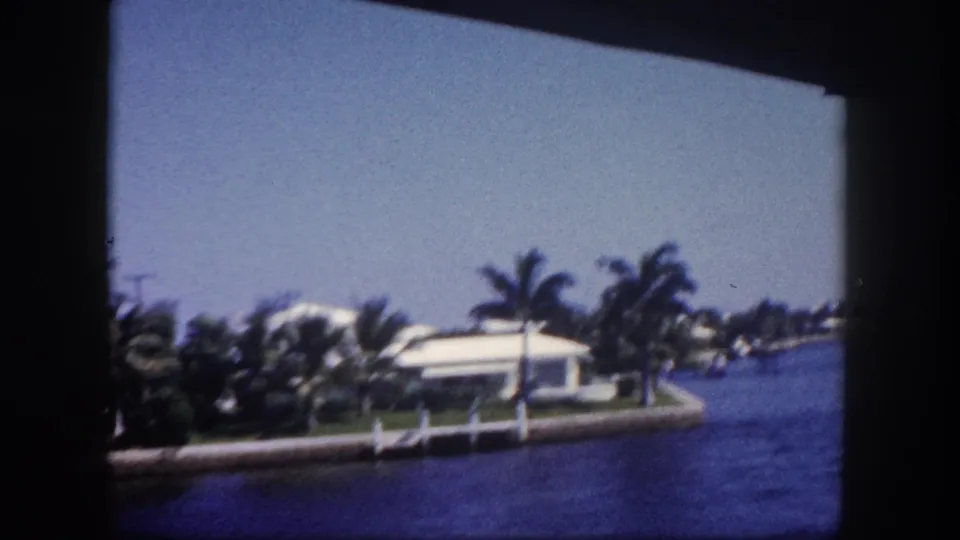 1960 Tampa home-movie frame with a low white waterfront house, palms, seawall posts, and blue canal water near the edge of the film frame.