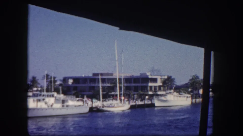 1960 Tampa channel scene with sailboats, dock pilings, palm trees, and a waterfront building in soft 8mm color.
