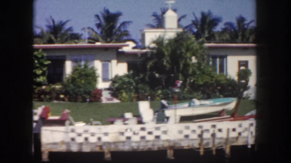 1960 Tampa-area home-movie frame showing a waterfront house, palm landscaping, decorative seawall, and small boat by the lawn.