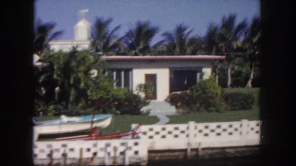 1960 Tampa-area waterfront residence with palm trees, hedges, a small boat, and patterned seawall viewed from the water.