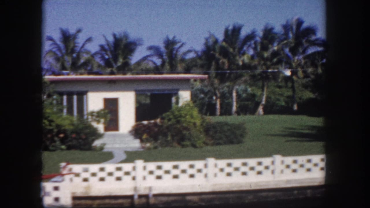 White stucco house, palm trees, lawn, patterned fence, and a small boat near the foreground.