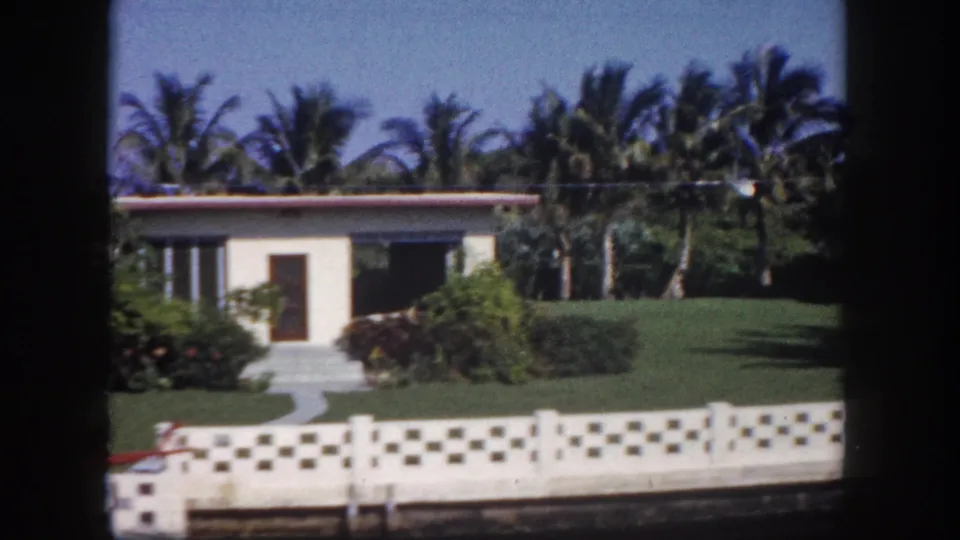 1960 Tampa home-movie frame showing a white stucco coastal house with red roof, palm trees, lawn, white patterned fence, and small boat.