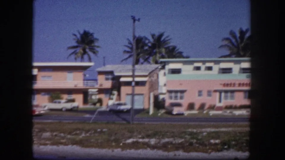 Low-rise Tampa roadside buildings, including a pink facade, sit beside a road with palms, parked cars, utility poles, and sandy ground.