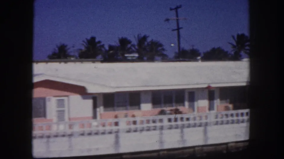 1960 Tampa frame of a pastel single-story home behind a low white fence, with palm trees and a utility pole against blue sky.