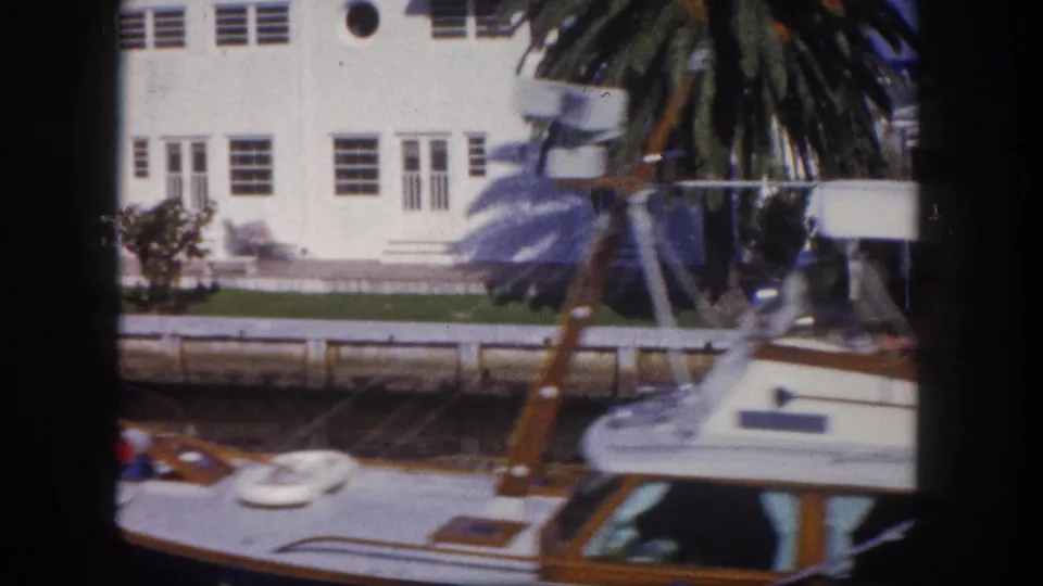 1960 Tampa 8mm frame with a white waterfront building, palm, seawall, canal water, and partial small boat in the foreground.
