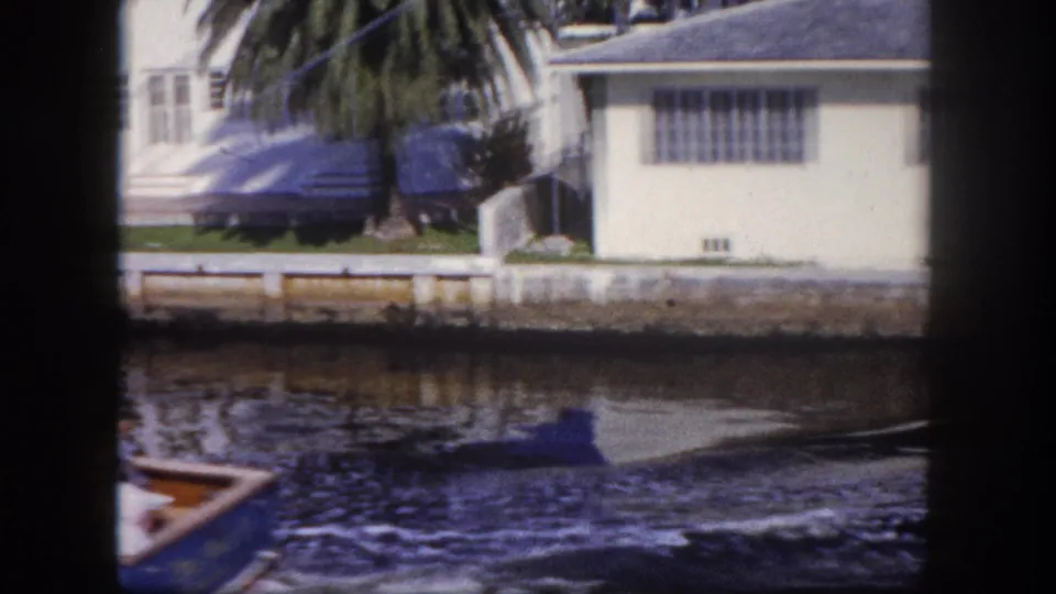 1960 Tampa waterfront frame showing a small boat on canal water near a white building, palm tree, seawall, and lawn.