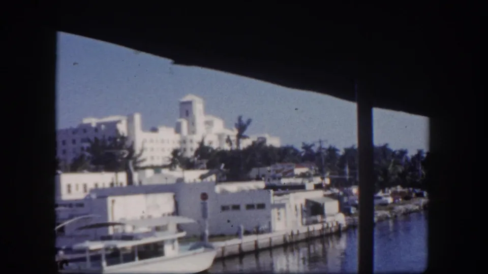Bright 1960 Tampa waterfront scene with moored boats, a concrete edge, white buildings, and palms beyond the water.