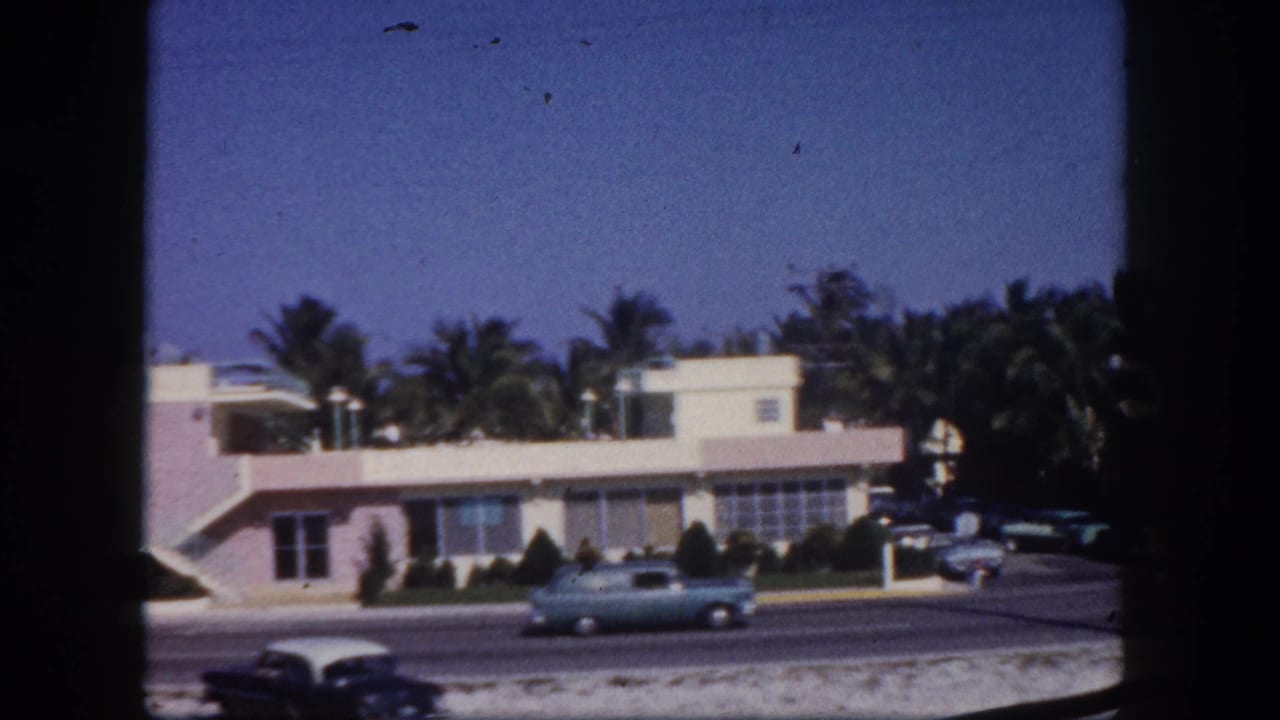 Street with a light-colored car passing low modern buildings and palm trees.