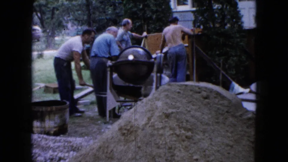 In Wauconda, Illinois, several adult men work around a portable cement mixer with a large sand pile in the foreground.