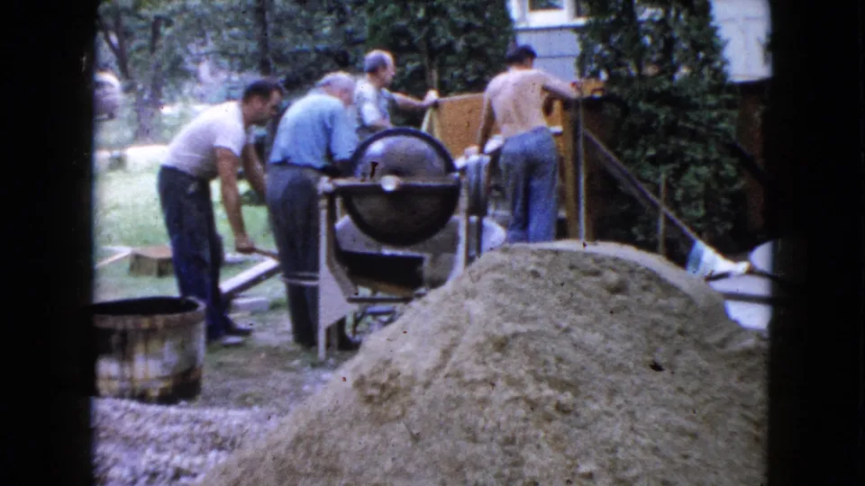 Adult men in casual clothing work around a cement mixer and chute in a Wauconda yard, with sand piled close to the camera.