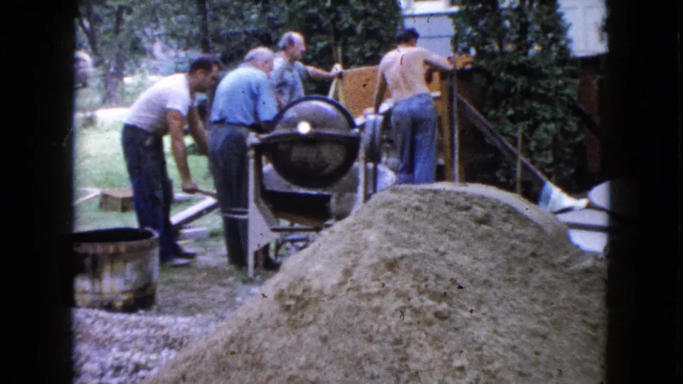 A shirtless adult man and others work near a cement mixer by house steps in the supplied 1960 Wauconda home-movie scene.