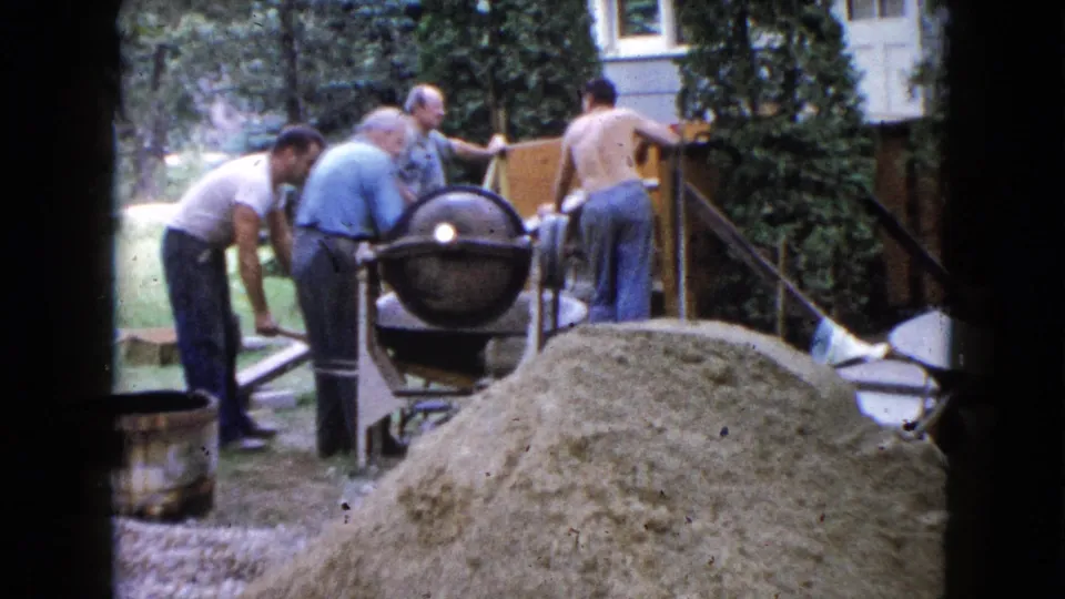 Four men appear to continue foundation work in a Wauconda backyard, with a cement mixer, sand pile, steps, and trees visible.