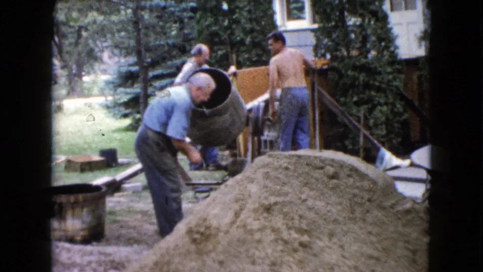 1960 Wauconda, Illinois frame with a man leaning near the mixer as other adult men handle porch construction materials in the backyard.