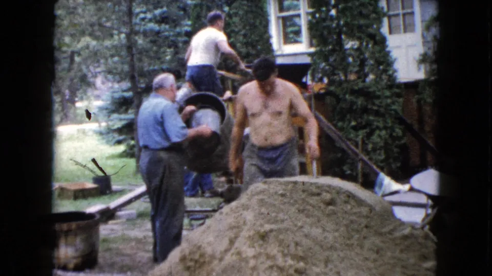1960 Wauconda, Illinois frame showing a shirtless man near the porch while another man manages the mixer beside a large sand pile.