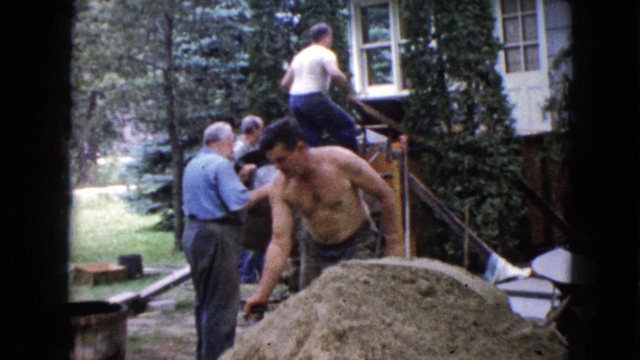 In 1960 Wauconda, Illinois footage, adult men work near house steps while a shirtless man handles material beside a sand or gravel pile.