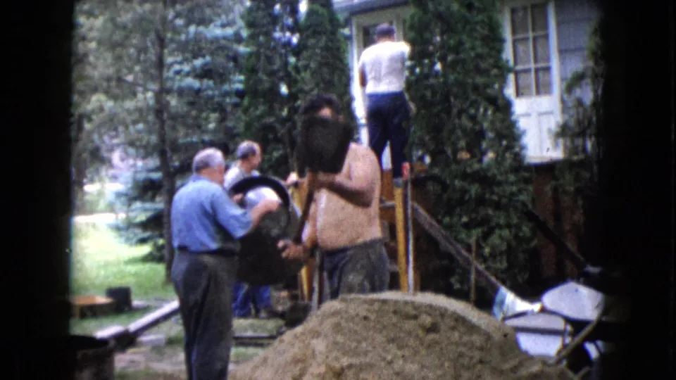 A shirtless worker lifts or tips a dark pail near a mound of material while other adult men continue work by the house steps.