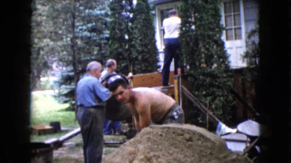 In the yard work scene, the shirtless man bends beside the sand or gravel pile as other men remain near the house steps and ladder.