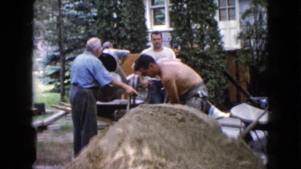 1960 Wauconda 8mm frame of men working with a cement mixer and sand pile outside a residential house.