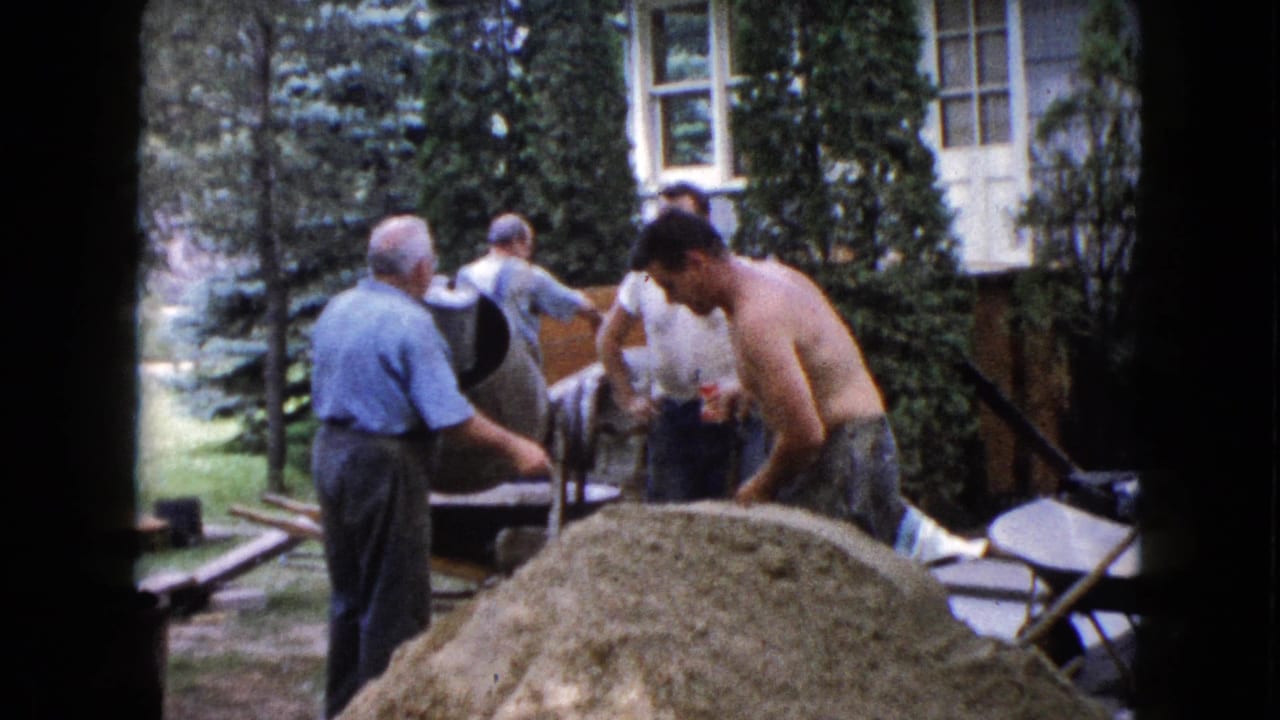 Men work around a sand pile, concrete mixer, and wheelbarrows in a backyard.