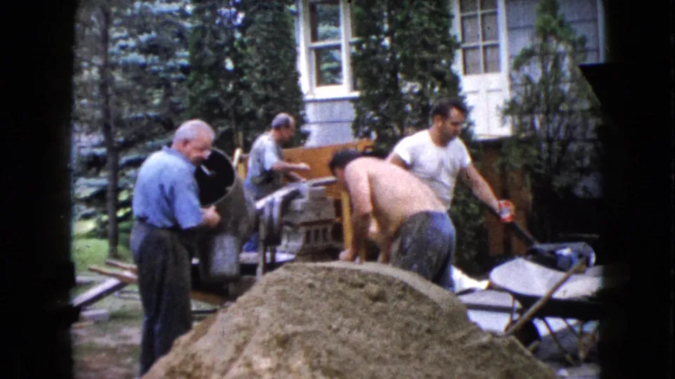 Adult men continue backyard concrete work in Wauconda, Illinois, with a mixer, wheelbarrows, and a house visible behind them.
