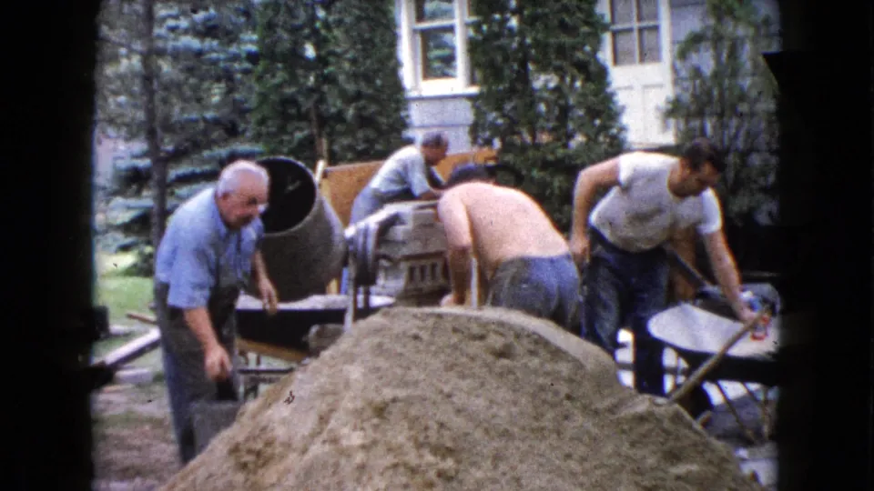 A 1960 Wauconda backyard scene shows men handling concrete materials near a mixer, wheelbarrows, sand pile, and house exterior.