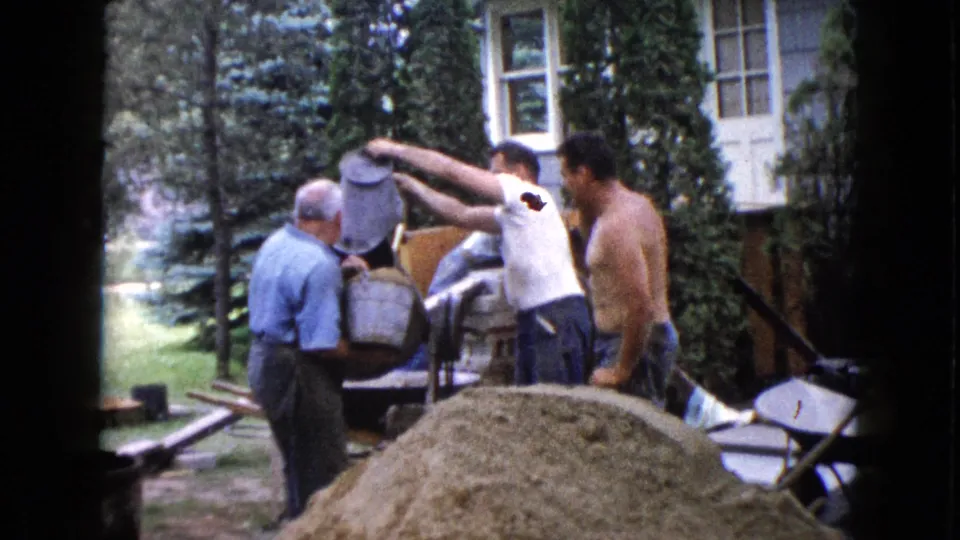 One man raises a bucket while two others stand near the mixer and sand pile during the 1960 backyard concrete preparation scene.