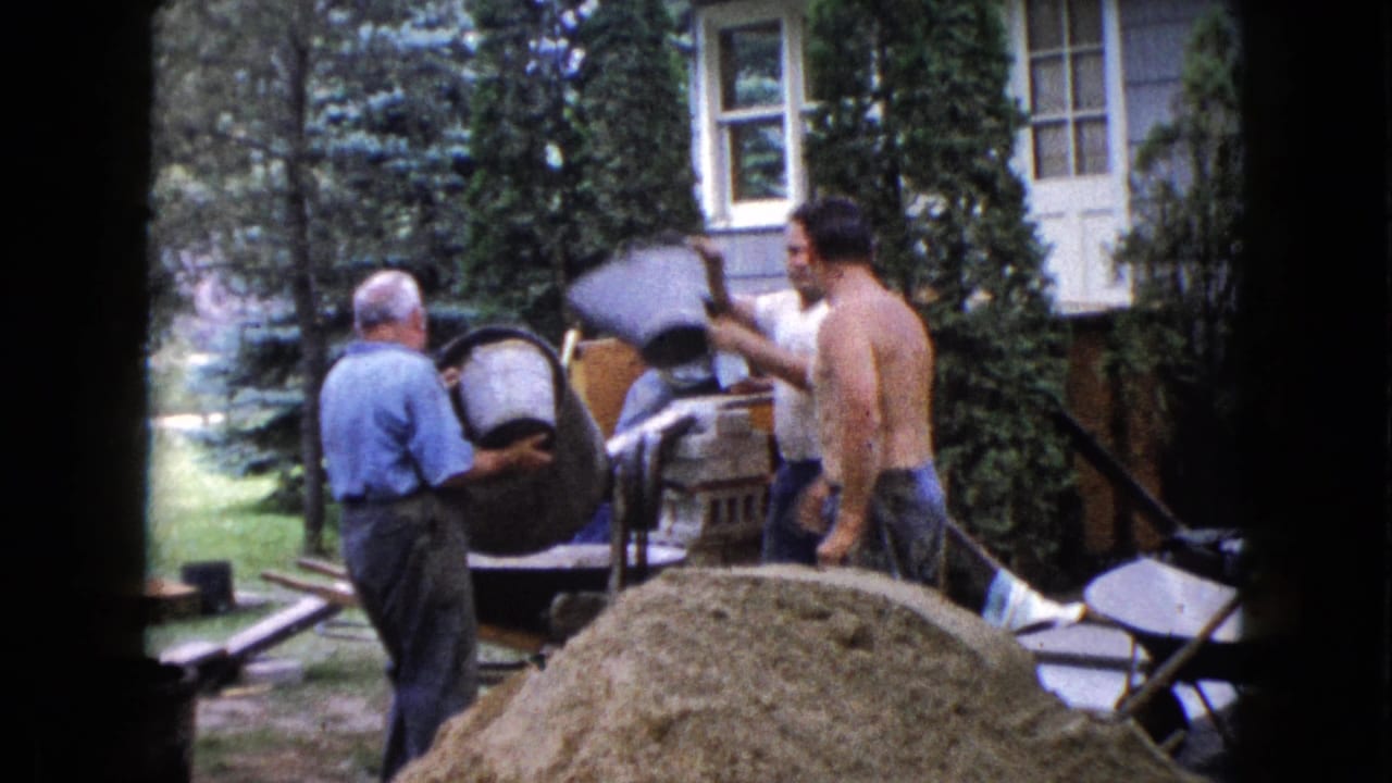 1960 Wauconda home-movie frame showing adult men working around a cement mixer beside a large sand pile in a suburban yard.
