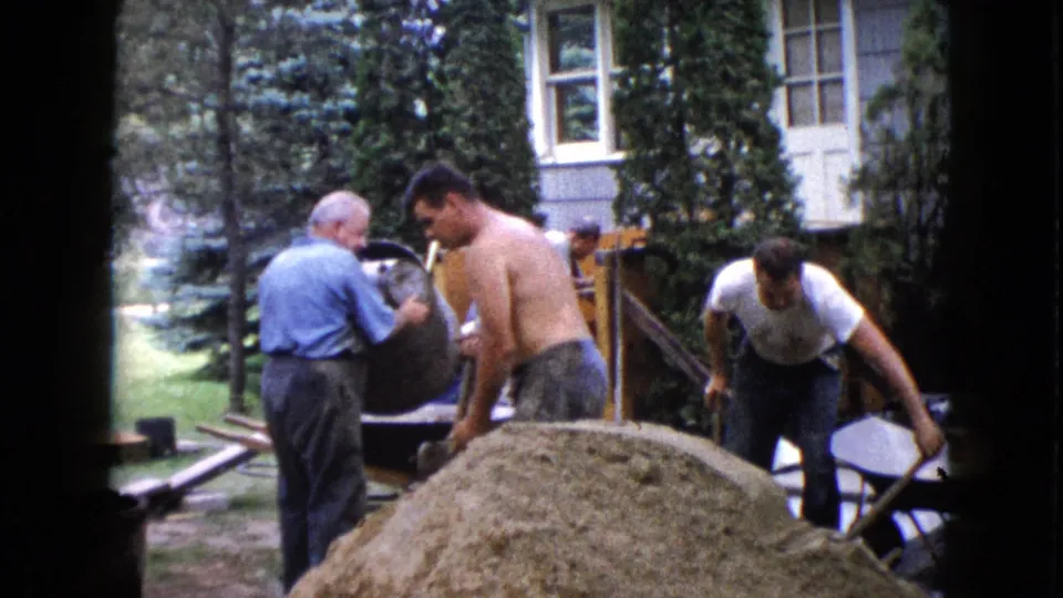 1960 Wauconda frame showing men in a residential yard working with a cement mixer, sand pile, and hand tools.
